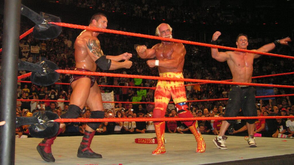 John Cena with Bautista and Hulk Hogan in the WWE wrestling ring. They appear to be celebrating with an equally enthusiastic crowd visible in the background.