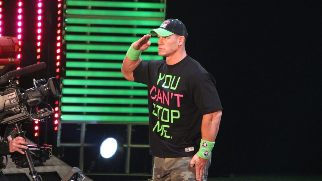 John Cena in his "You Can't See Me" t-shirt giving a military salute. A professional shooting camera set is visible on the left side.