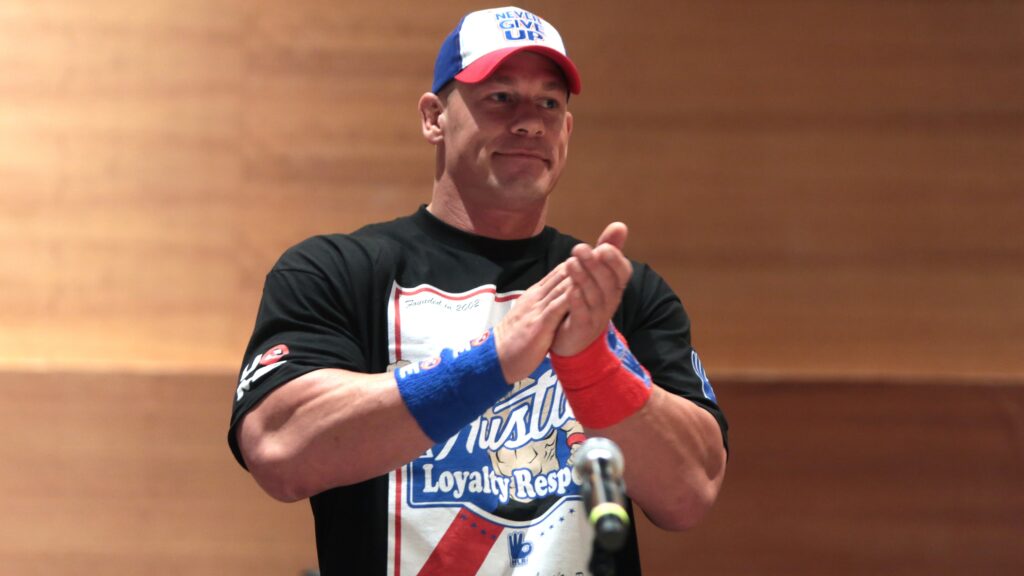 John Cena in a red, white and blue "Never Give Up" cap and "Hustle Loyalty Respect" t-shirt, clapping. There's a blurred microphone in the foreground.