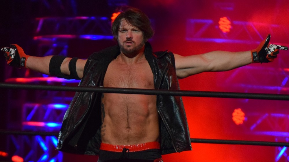 AJ Styles in a black leather jacket with arms outstretched in the WWE ring. The background is lit with red and blue light.