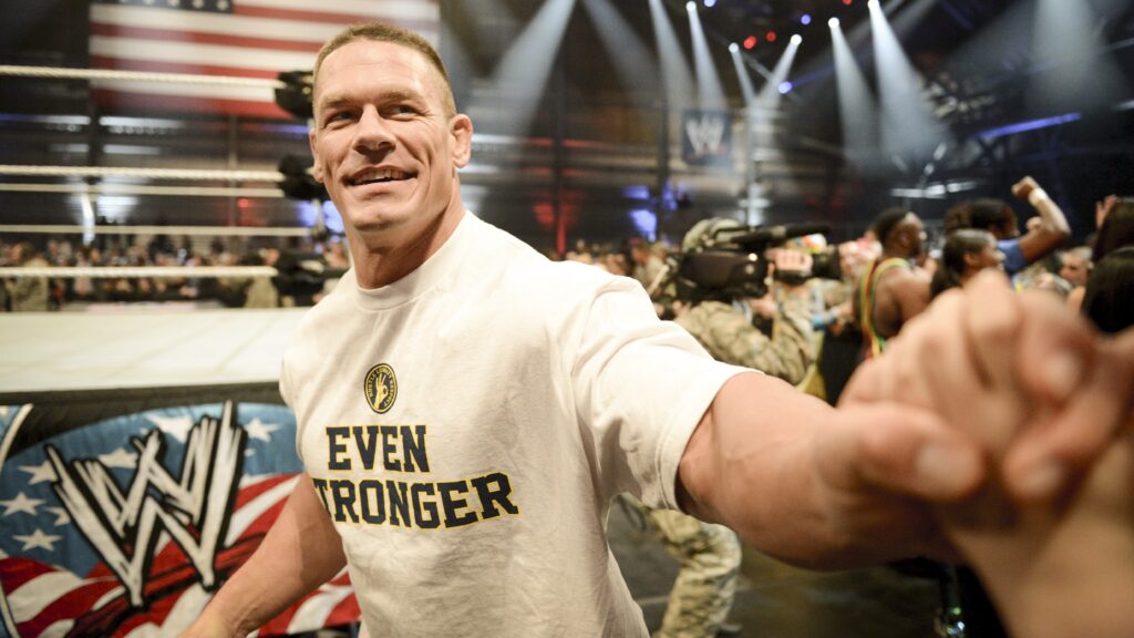 John Cena in a white "Even Stronger" t-shirt smiling and reaching to hold a fan's hand while at a WWE event.