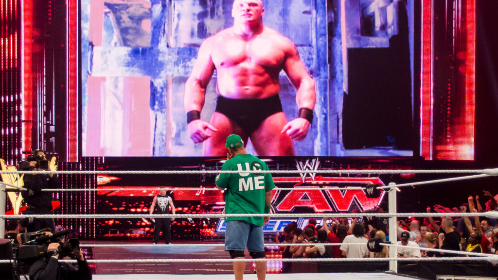 John Cena in his iconic green gear confronting Brock Lesnar on the entrance ramp with a video screen showing Lesnar's image in the background.