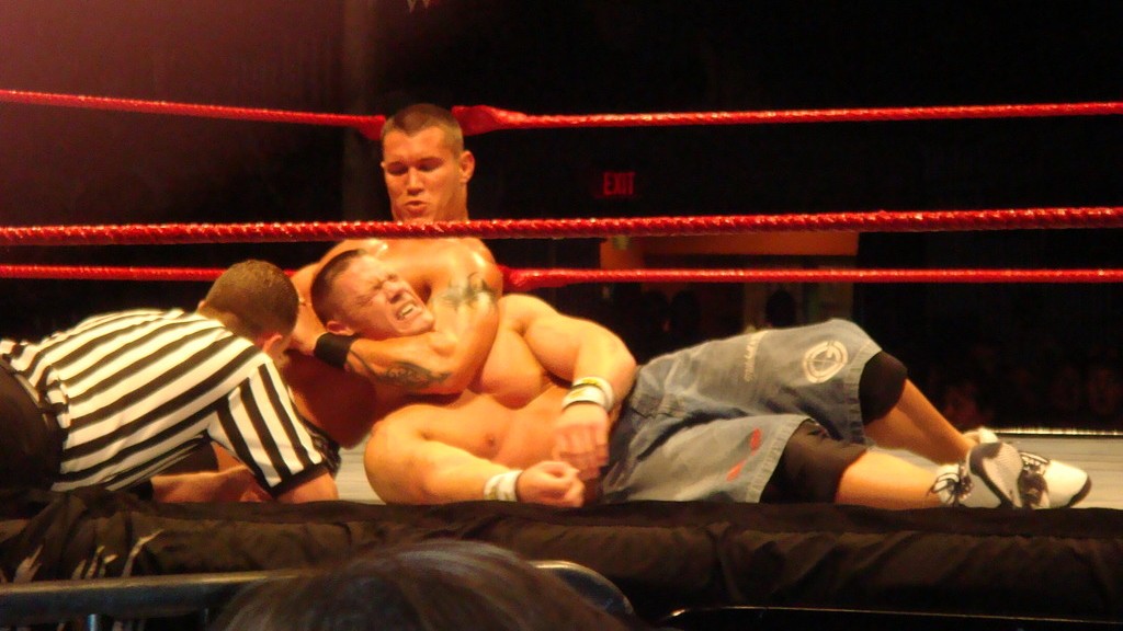 Randy Orton choke-holds John Cena during a WWE match as the referee watches closely.