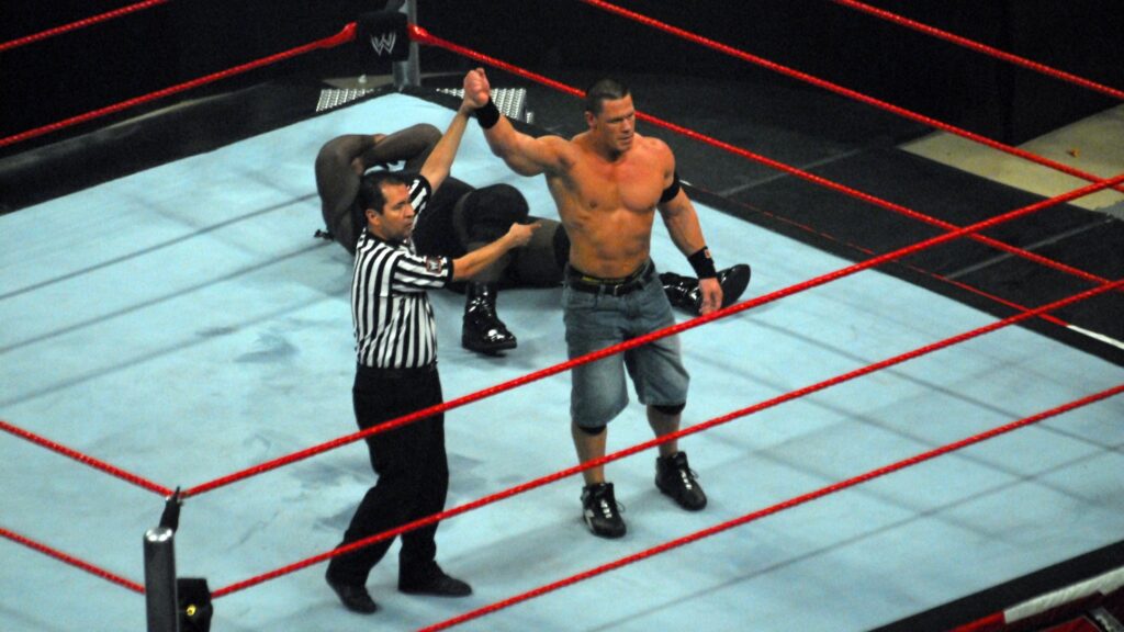 John Cena in jean shorts being declared the winner of a WWE match, with a referee raising his arm while his opponent, Mark Henry, lies on the mat.