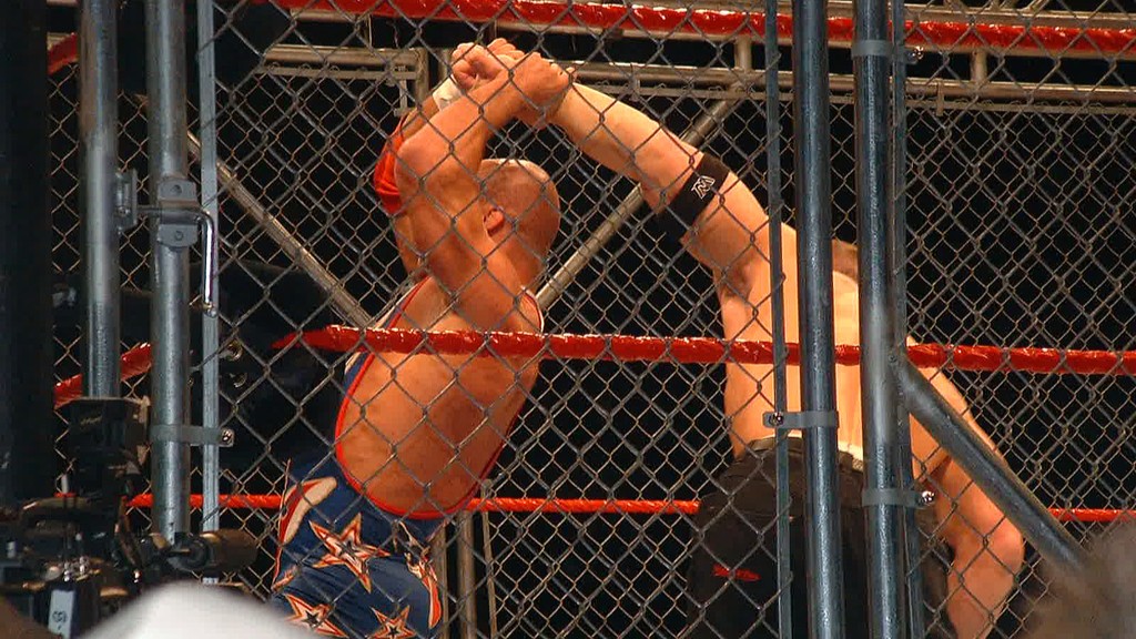 Kurt Angle and John Cena fighting inside a steel cage with Angle holding Cena's arm over his head.