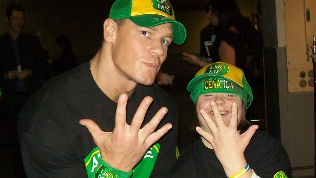 John Cena posing with wish child Savannah who is wearing Cena's merchandise including a green "CENATION" cap. Both are making Cena's iconic "You Can't See Me" hand gesture with their fingers spread in front of their faces.