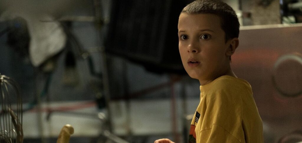 Millie Bobby Brown as Eleven in a still from Stranger Things, looking shocked during an intense moment.