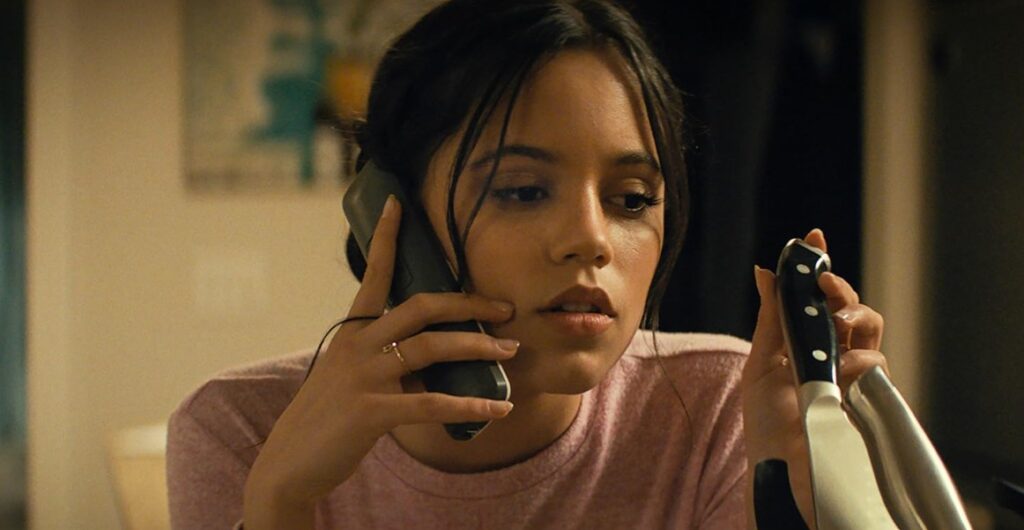 Jenna Ortega as Tara Carpenter in Scream VI, is talking on the phone while holding a knife.