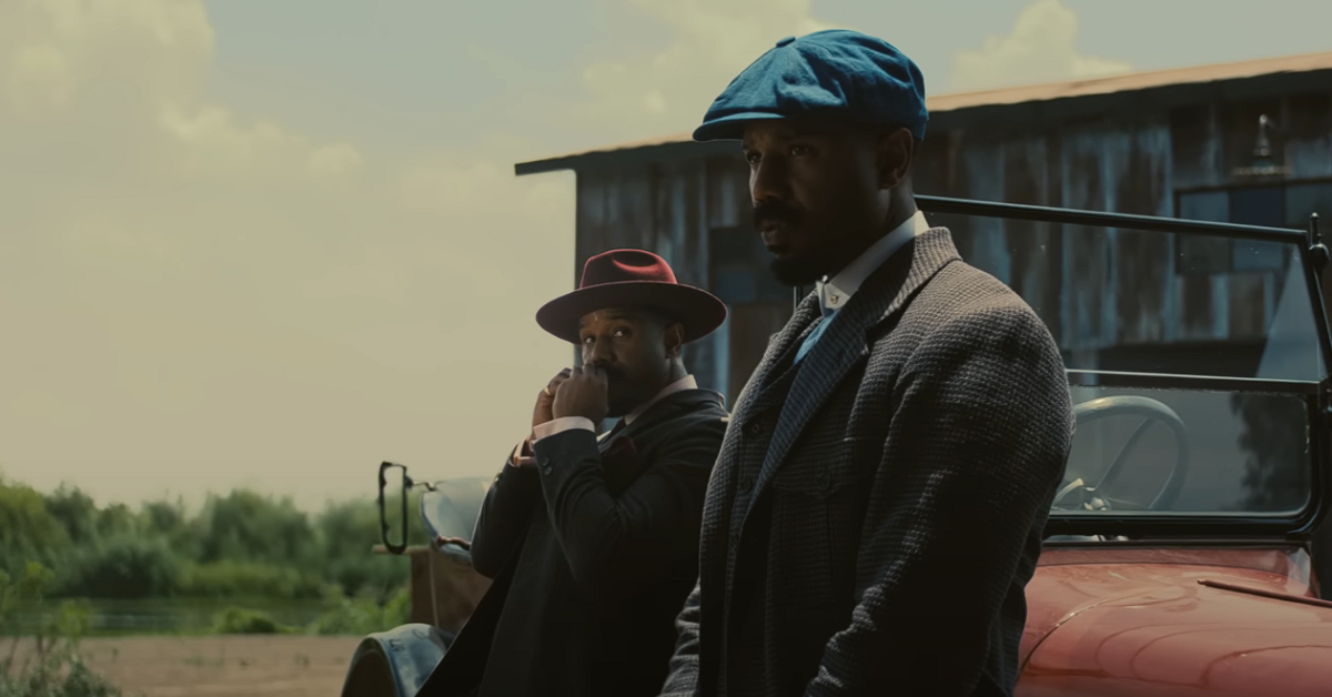 Two Michael B Jordan in a blue and red hat leaning against a car. 