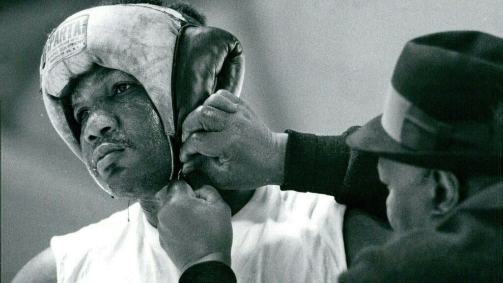 A black and white photo of George Foreman having his boxing headgear adjusted before a fight.