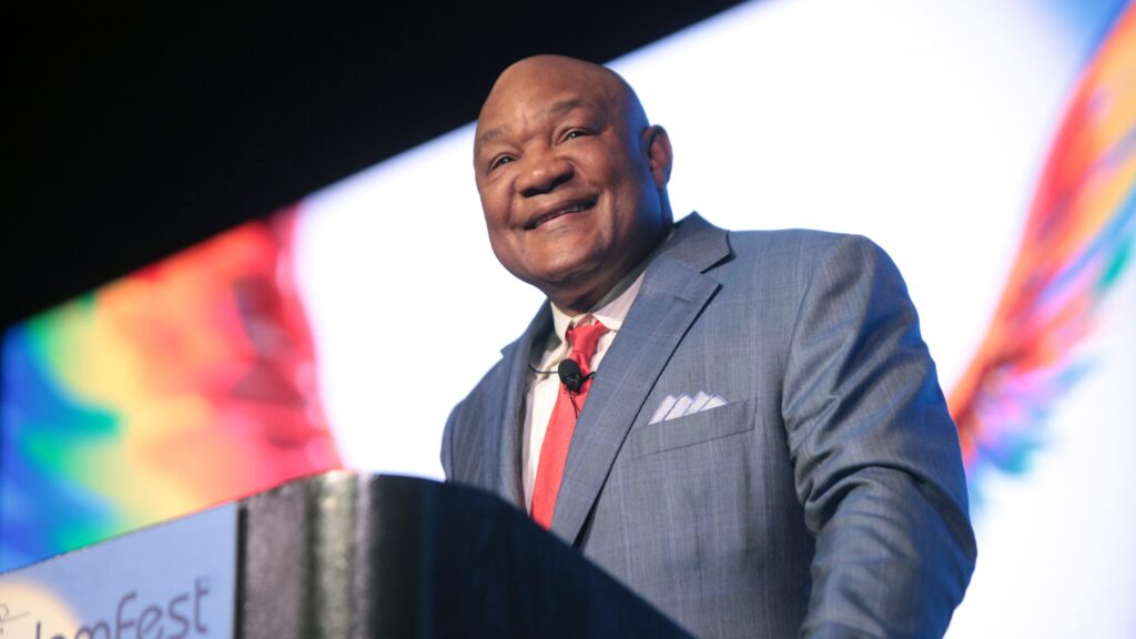 George Foreman in a light blue suit with a red tie, speaking at what appears to be a conference or event.