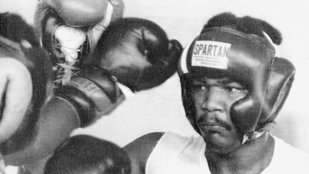 A black and white photo of a young George Foreman in boxing headgear during training or an amateur match.