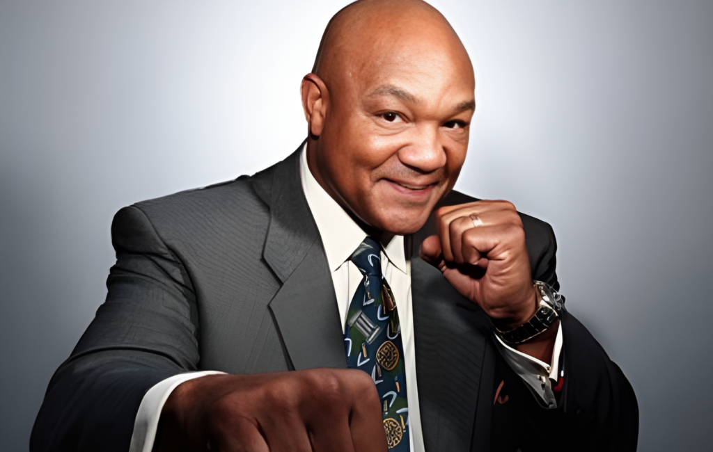 A professional portrait of George Foreman in a dark suit with a patterned tie. He's posing with one fist raised with a warm smile on his face.