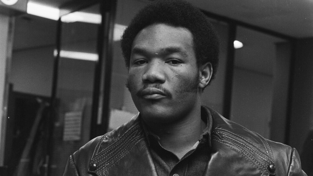 A black and white portrait photograph of a young George Foreman in a leather jacket. He's looking directly at the camera with a serious expression.