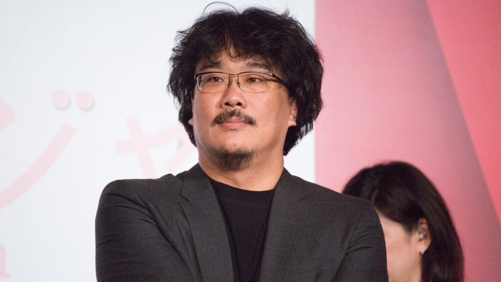 10 Most Successful Non-American Directors in Hollywood 10 Bong Joon-ho smiling during Okja Japan Premiere. He has a thoughtful expression and is photographed against a light background with a pinkish tint.