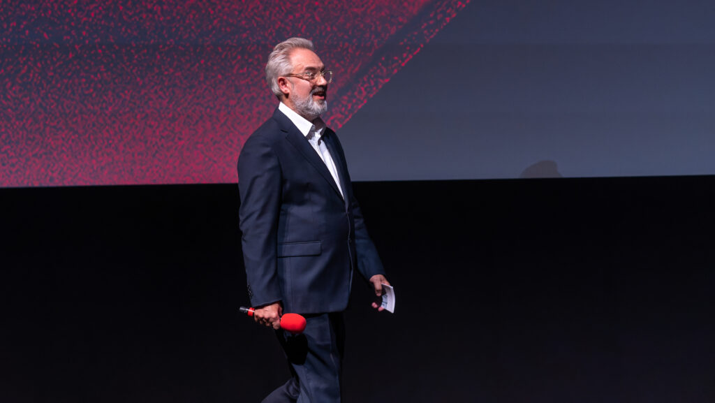 10 Most Successful Non-American Directors in Hollywood 13 Sam Mendes, wearing a dark suit is speaking at an event, holding a red microphone in his hand. He is on the BFI LFF Gala - Royal Festival Hall stage.