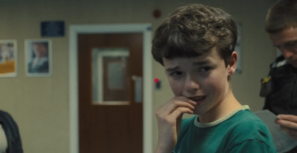 Owen Cooper as Jamie Miller in the series Adolescence crying as an officer behind him checks papers.