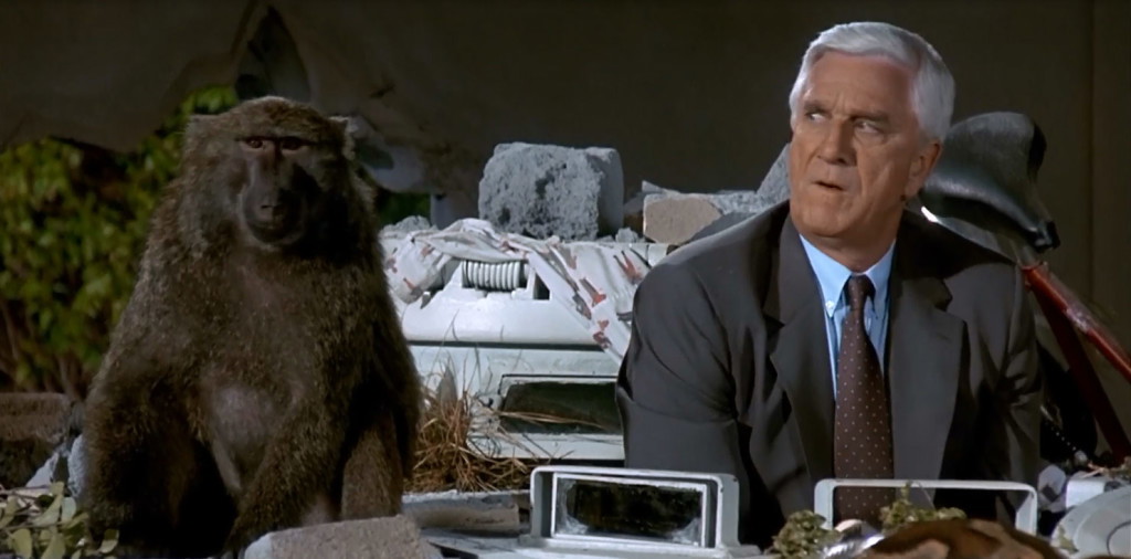 ‘The Naked Gun’ Trailer: Liam Neeson Proves David Zucker Shouldn’t Compare It To Internet’s Most Infamous Video 3 Leslie Nielsen sitting and looking suspiciously to his right side where a monkey is sitting.