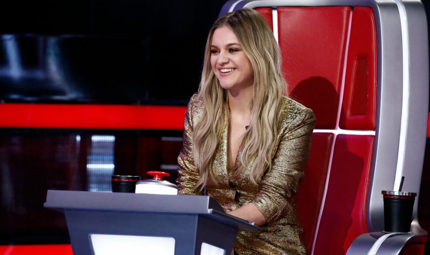 Kelsea Ballerini, sitting in the red judge's chair in the reality show The Voice with a smile on her face.