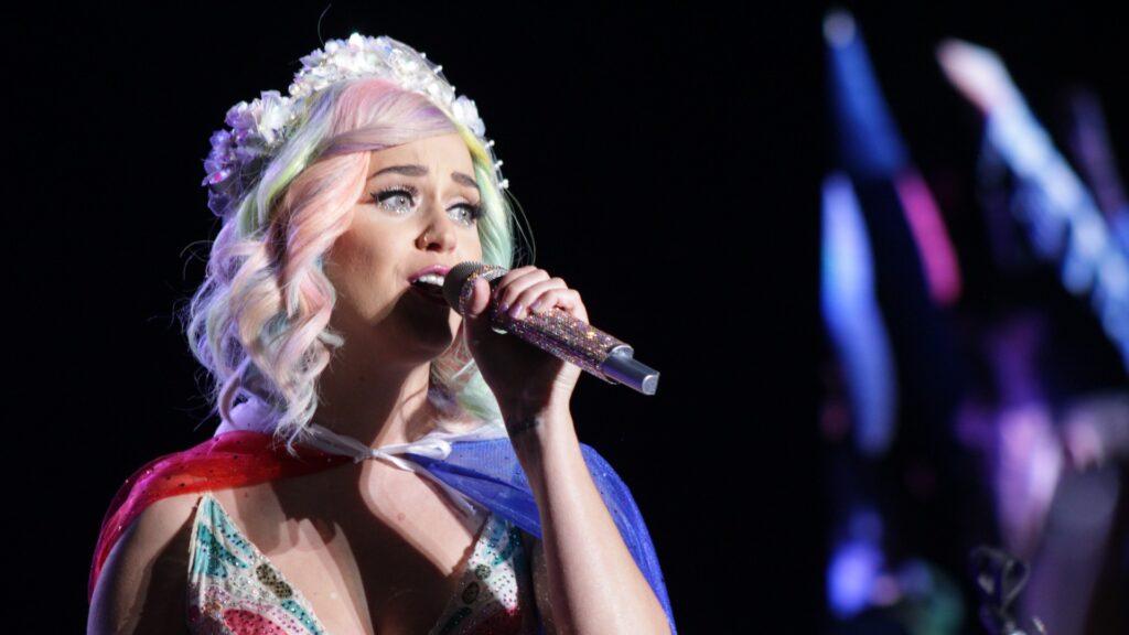 Katy Perry singing with a bedazzled microphone, wearing a floral crown and featuring pastel-colored hair with pink, lavender and mint green tones during The Prismatic World Tour in Chile. She's in a colorful outfit with patriotic red, white and blue elements, performing against a dark background with stage lighting visible.