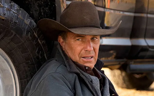 Kevin Costner wearing a hat and hiding behind a car in Yellowstone