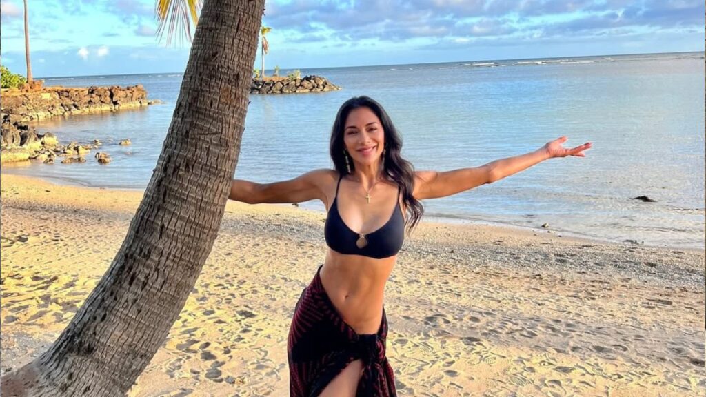 Nicole Scherzinger spreads her arms wide in a two -part piece with the backdrop of a beach