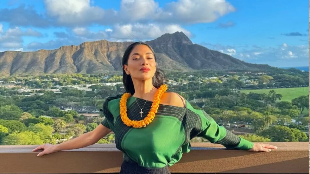 Against the background of a picturesque mountain, Nicole Scherzinger leans onto the balcony