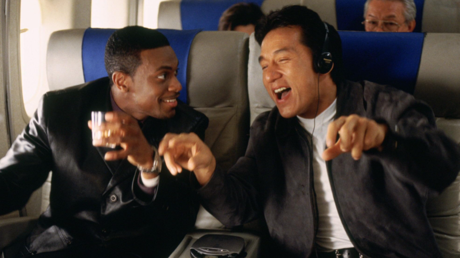 Lee and Carter are on an airplane. Lee has headphones on and appears to be grooving to the music. Beside him, Carter is holding a glass and smiling at Lee.