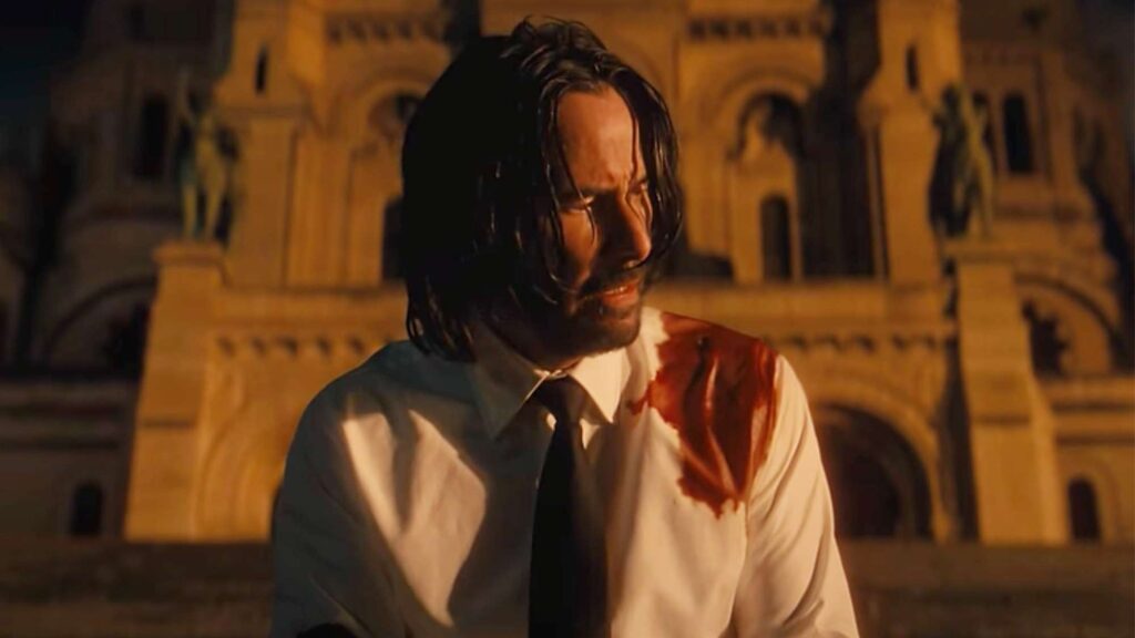 John Wick 5: Everything We Know So Far 1 Keanu Reeves as John Wick in a white dress shirt with blood stains, looking towards his side with a concerned or confused expression.