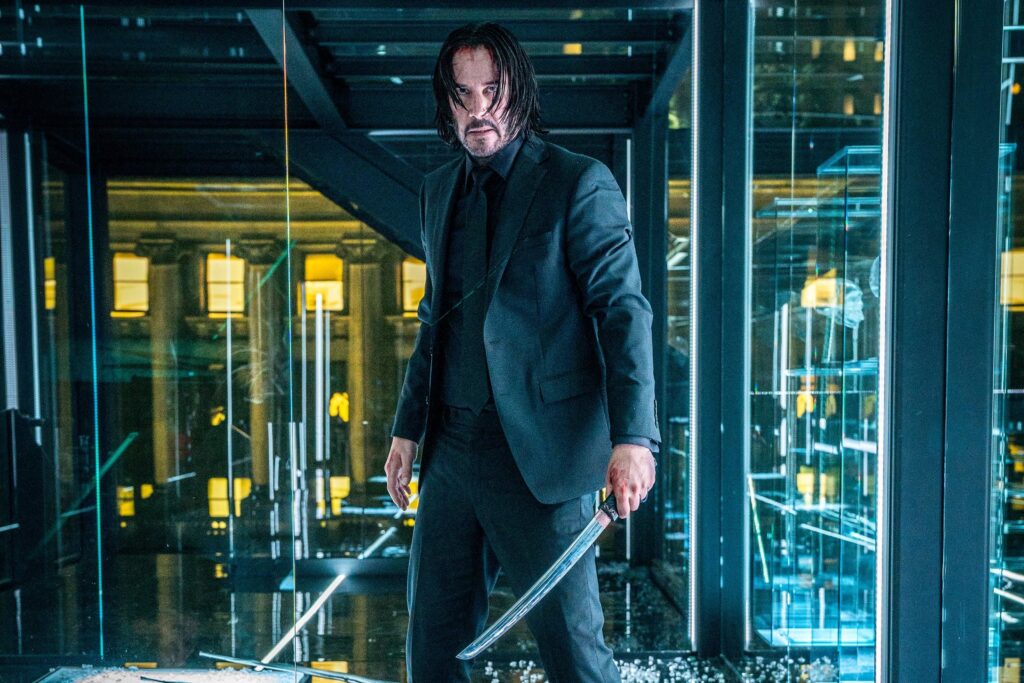 John Wick 5: Everything We Know So Far 2 Keanu Reeves as John Wick in his signature black suit holding a large knife, standing in a modern glass office building with reflective surfaces and yellow lighting.