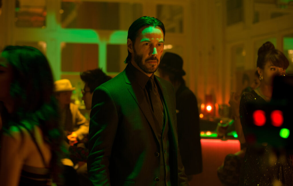 John Wick 5: Everything We Know So Far 4 Keanu Reeves as John Wick in a dark suit standing in a nightclub setting with green and red neon lighting, surrounded by blurred figures in the background.