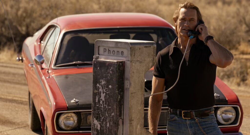 Josh Holloway by the pay phone talking to someone while a red car is parked behind him