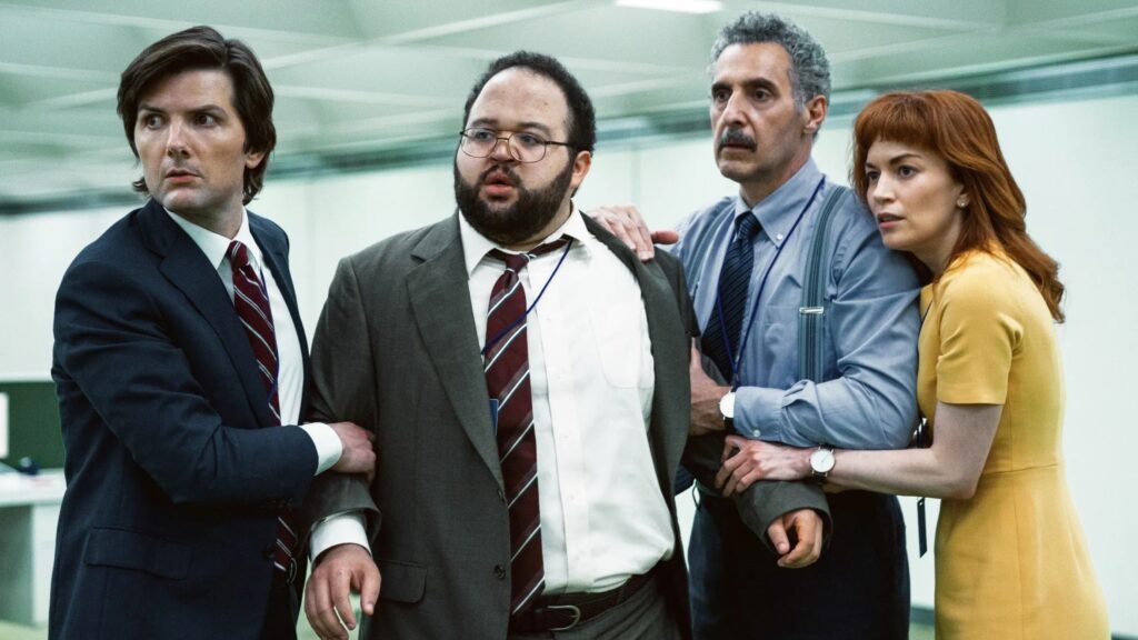 20 Absolutely Best TV Shows to Watch Right Now (June 2025) 51 Mark (Adam Scott), Dylan (Zach Cherry), Irving (John Turturro), and Helly (Britt Lower) in business attire stand together in a modern office corridor.