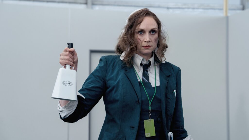 20 Absolutely Best TV Shows to Watch Right Now (June 2025) 53 Lorne (Gwendoline Christie) wearing a dark teal business suit and green lanyard holds up what appears to be a white electronic device.