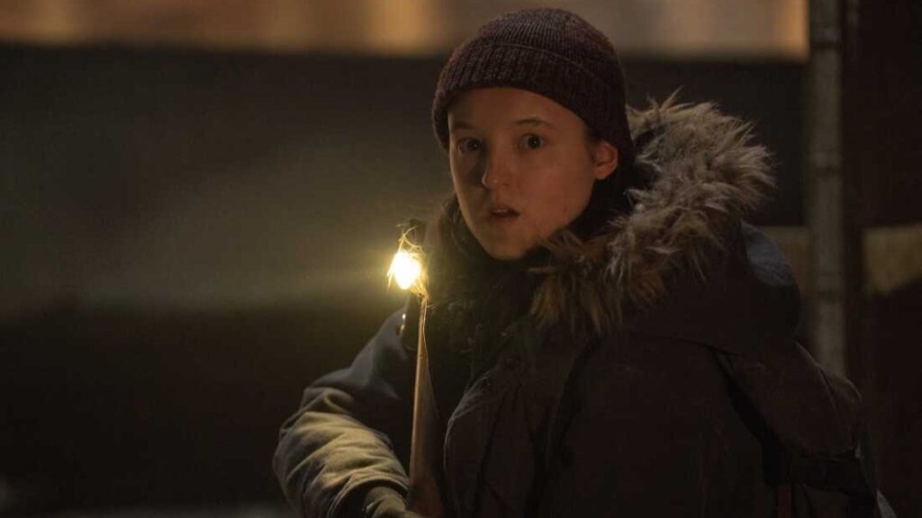 20 Absolutely Best TV Shows to Watch Right Now (June 2025) 58 Ellie wearing a dark knit cap and winter jacket with fur-lined hood holds a flashlight in a dimly lit indoor setting.