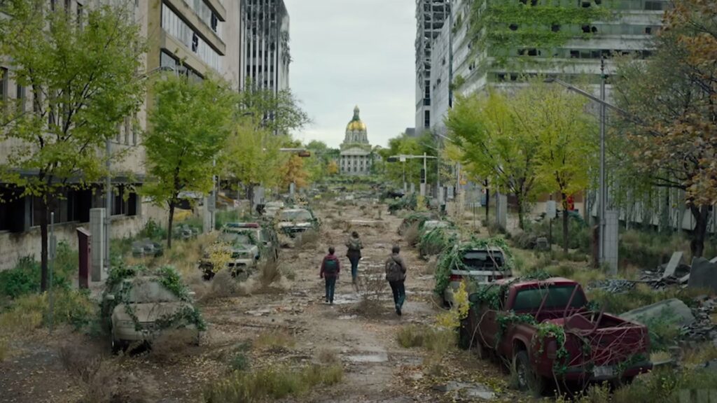 20 Absolutely Best TV Shows to Watch Right Now (June 2025) 57 A wide shot of a destroyed city street with abandoned cars, rubble, and overgrown vegetation, while three small figures walk down the debris-filled road toward a domed government building visible in the distance.