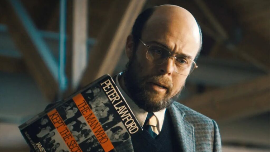 20 Absolutely Best TV Shows to Watch Right Now (June 2025) 74 Trent (Ian Merrigan) wearing glasses and a dark suit holds a book by Peter Lanford title "The Man Who Kept the Secret" in a dimly lit, industrial-looking setting.
