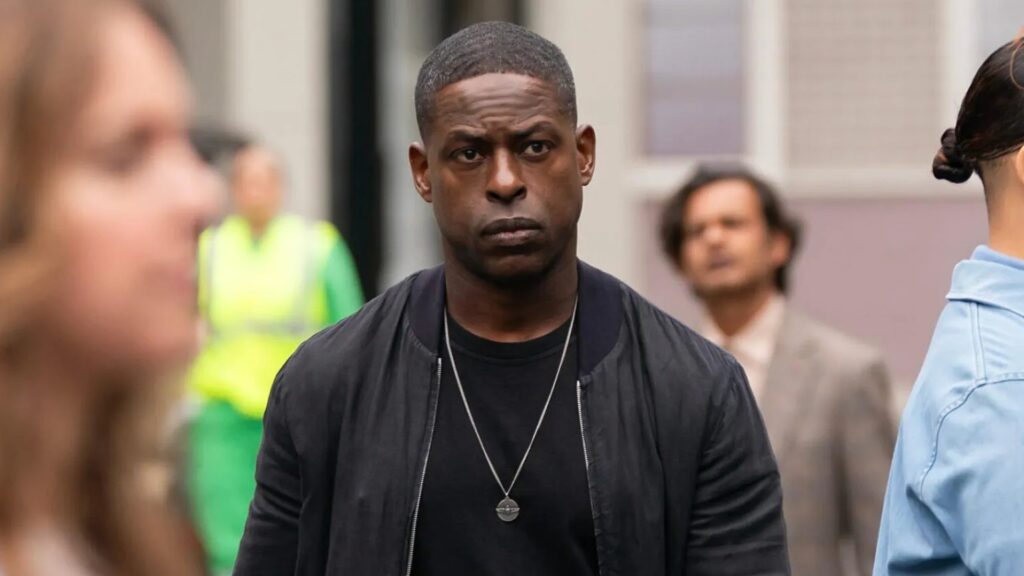 20 Absolutely Best TV Shows to Watch Right Now (June 2025) 75 Xavier Collins wearing a dark bomber jacket over a black t-shirt with a chain necklace walks on a city street with blurred figures and urban buildings visible in the background.