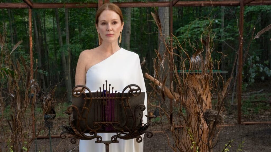 20 Absolutely Best TV Shows to Watch Right Now (June 2025) 81 Michaela Kell (Julianne Moore) in an elegant white one-shoulder dress stands behind an ornate decorative podium in an outdoor garden setting with lush greenery in the background.