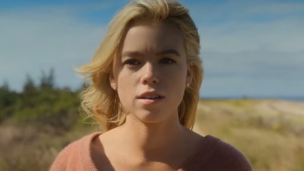 20 Absolutely Best TV Shows to Watch Right Now (June 2025) 83 Simone DeWitt (Milly Alcock) in a coral-colored sweater looks directly at the camera with a serious expression, positioned outdoors with a blurred natural landscape background.