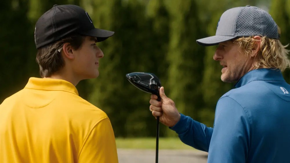 20 Absolutely Best TV Shows to Watch Right Now (June 2025) 87 Santi (Peter Dager) and Pryce Cahill (Owen Wilson) on a golf course - Santi in a yellow polo shirt and Cahill in blue golf attire with a cap, holding a golf club.