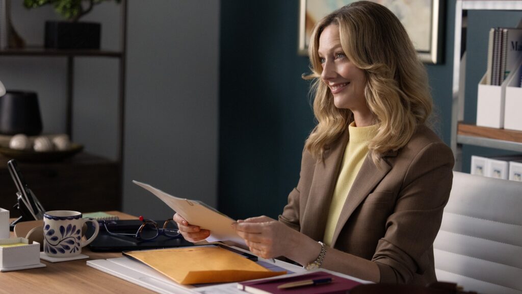 20 Absolutely Best TV Shows to Watch Right Now (June 2025) 89 Judy Greer (Amber-Linn) in a brown blazer and yellow top sits at a desk reading documents in a professional office setting.