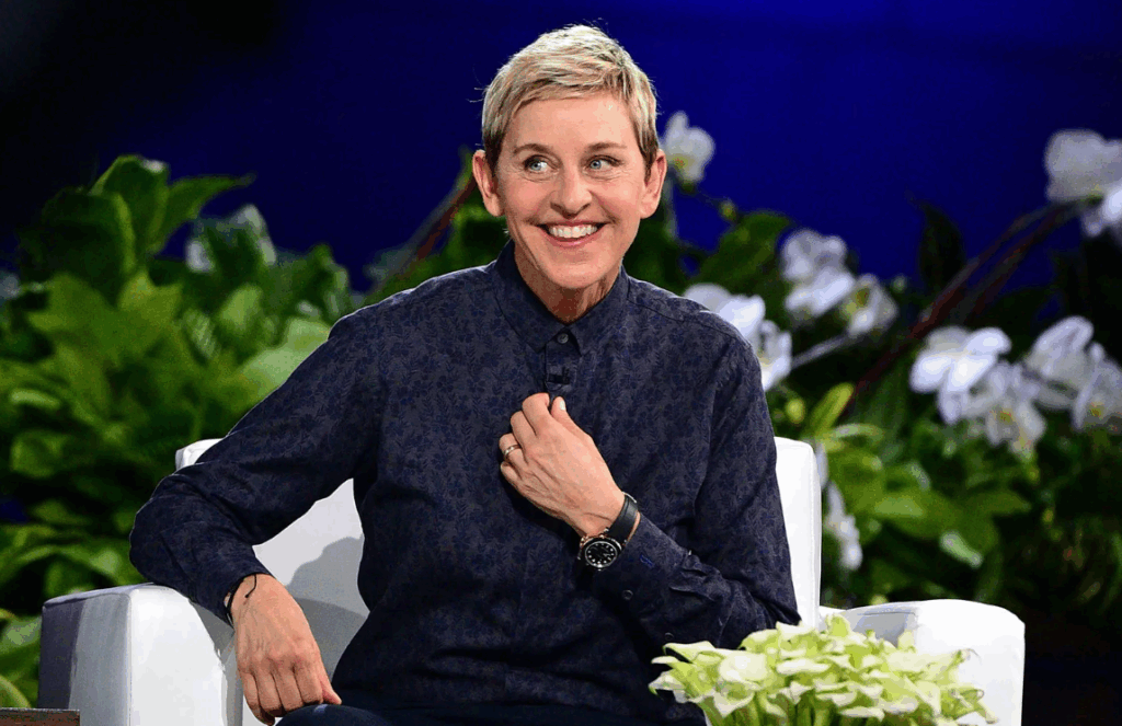 P Diddy’s Decades Long Friendship With Ellen Degeneres Explored 2 Ellen DeGeneres is sitting on a white chair in front of green plants and white flowers. She has a big smile on her face.