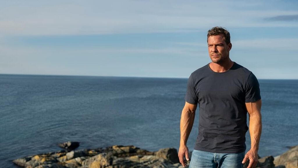 20 Absolutely Best TV Shows to Watch Right Now (June 2025) 91 Jack Reacher (Alan Ritchson) with dark hair wearing a black t-shirt stands on rocky coastline overlooking the ocean.