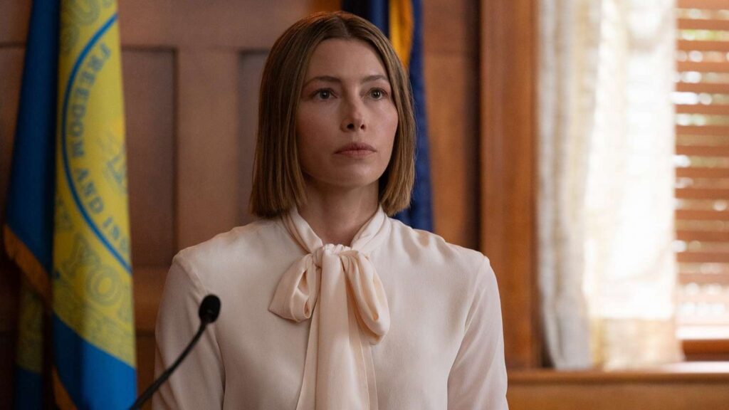 20 Absolutely Best TV Shows to Watch Right Now (June 2025) 98 Chloe Taylor (Jessica Biel) wearing a cream-colored blouse with a bow tie collar, at a podium with a microphone in a courthouse.
