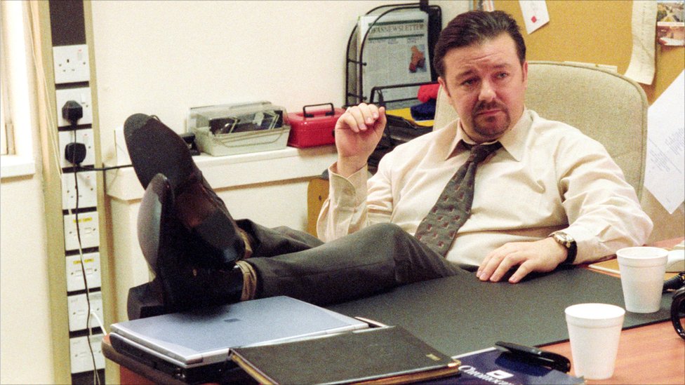 Ricky Gervais Says He and Steve Carell Were Just 2 Idiots Ad Libbing During His Cameo in The Office 2 Ricky Gervais' David Brent sitting with his feet up on the desk, The Office UK