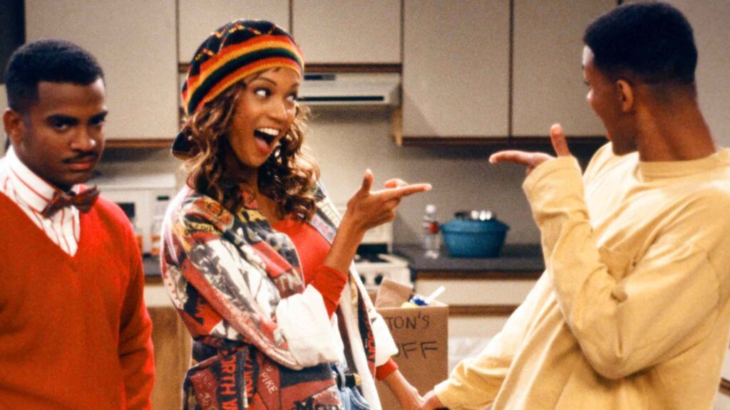 10 Insanely Popular TV Shows From the ‘90s You Can Stream on Netflix 13 Scene from The Fresh Prince of Bel-Air showing Tyra Banks in colorful '90s attire with a patterned headwrap, gesturing while speaking to Will in a kitchen setting.