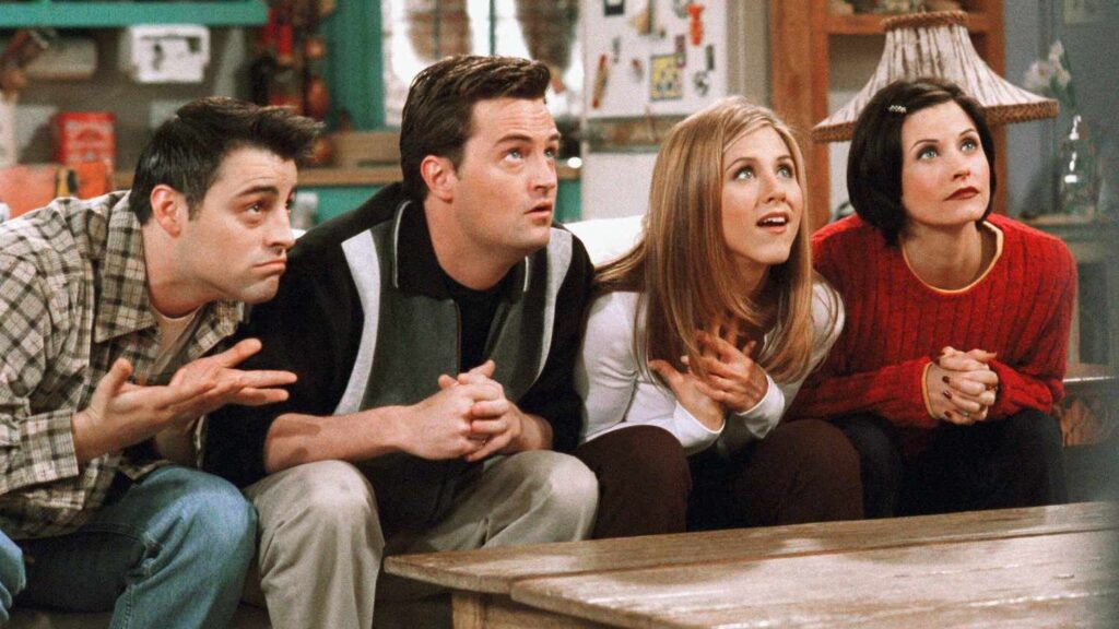 10 Insanely Popular TV Shows From the ‘90s You Can Stream on Netflix 17 Scene from Friends showing Joey, Chandler, Rachel, and Monica sitting together on a couch, all looking up with surprised expressions in Monica's apartment.