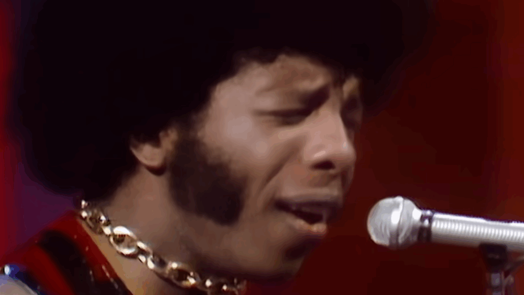 Sly Stone Dies at 82: What We Know About His Wife, Family, and Net Worth 7 Close-up of Sly Stone singing into a microphone, wearing a gold chain necklace against a reddish background on The Ed Sullivan Show.