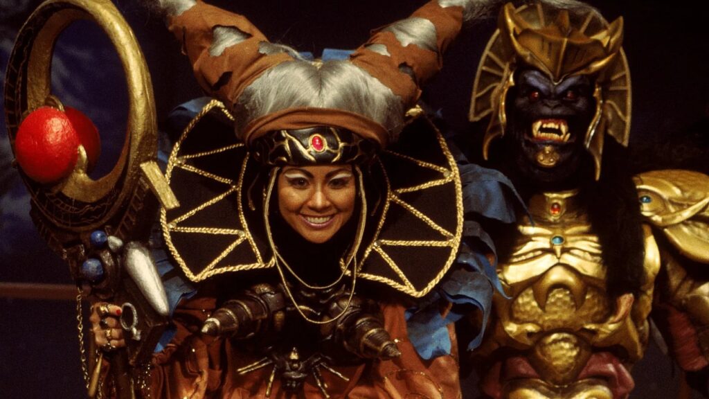 10 Insanely Popular TV Shows From the ‘90s You Can Stream on Netflix 28 Scene from "Mighty Morphin Power Rangers" showing Rita Repulsa in her elaborate golden and blue costume with ornate headdress, flanked by her monster minion in the background.